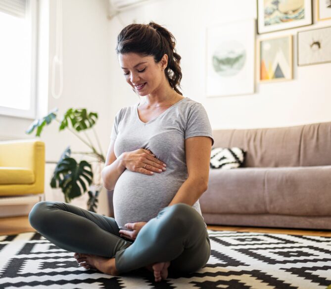 A pregnant woman sitting on the floor of her living room holding her baby bump representing can egg donation cause infertility.