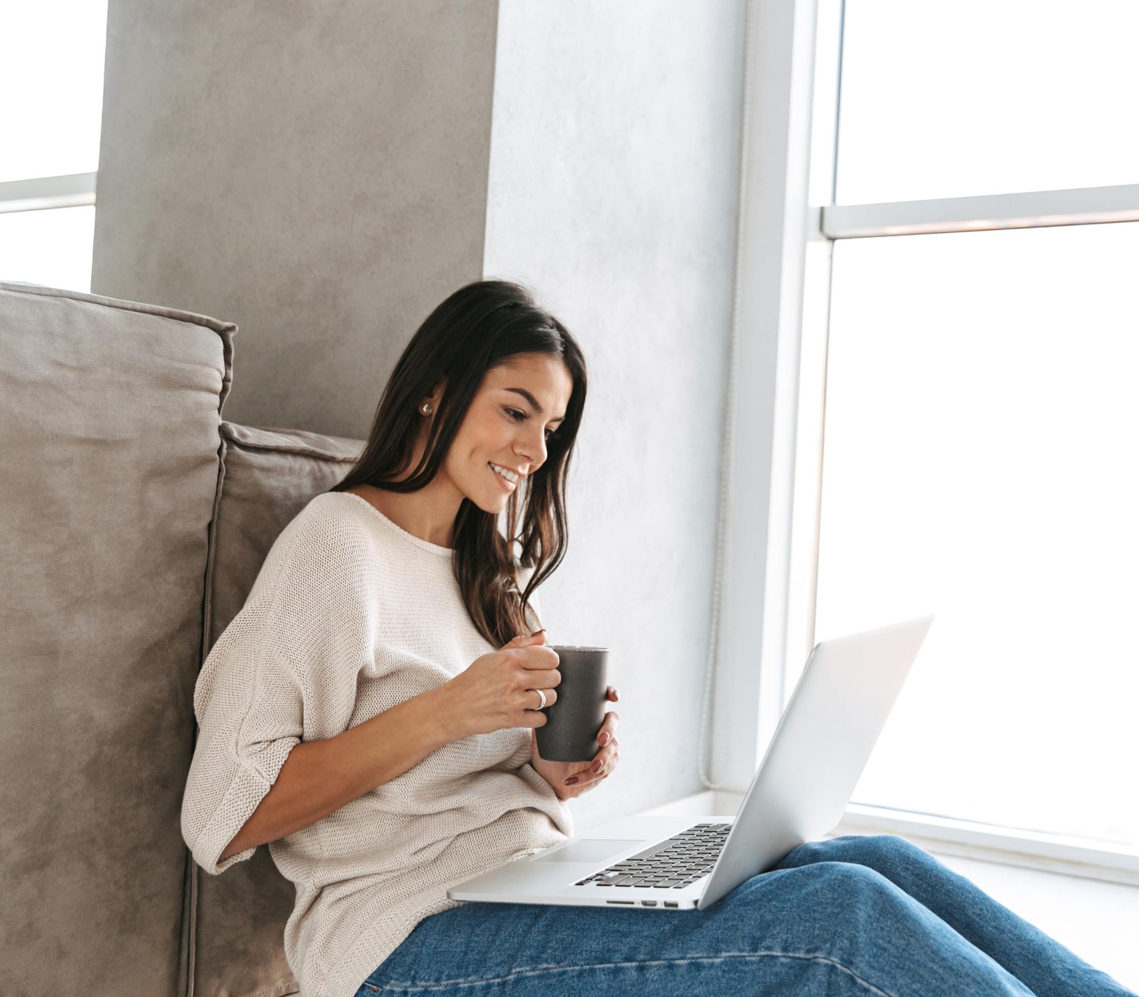 Women sitting on the floor with a cup of coffee, on her computer, smiling.